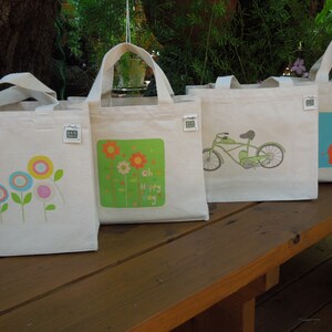 May include: Four white canvas tote bags with different designs. The first bag has a colorful flower design. The second bag has a green background with a flower design and the text "Oh Happy Day". The third bag has a green bicycle design. The fourth bag has a blue background with a red elephant design.