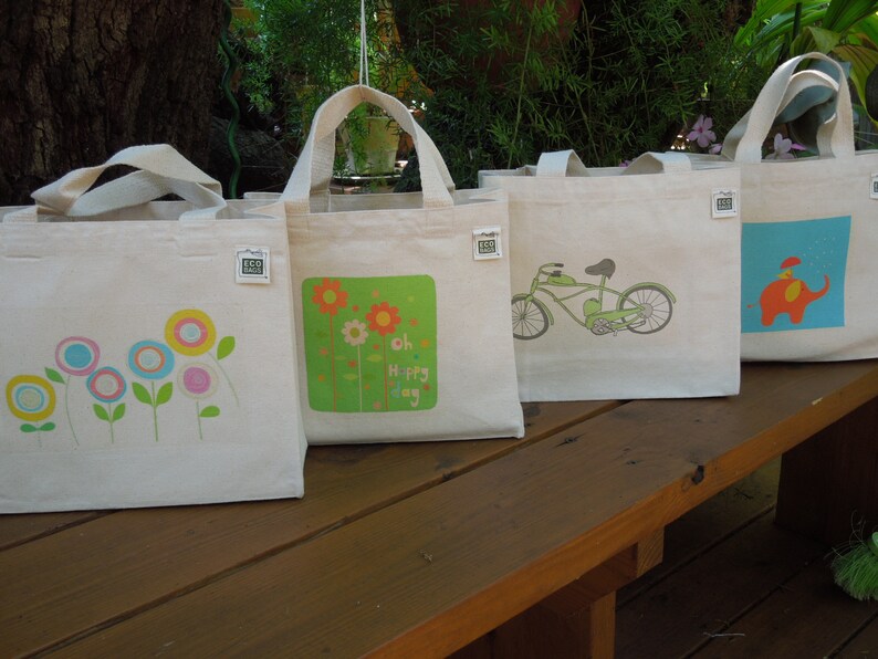 May include: Four white canvas tote bags with different designs. The first bag has a colorful flower design. The second bag has a green background with a flower design and the text "Oh Happy Day". The third bag has a green bicycle design. The fourth bag has a blue background with a red elephant design.