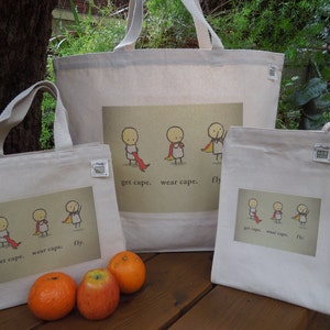 May include: Three white canvas tote bags with colourful illustrations of stick figures wearing capes. The text on the bags reads "get cape. wear cape. fly."