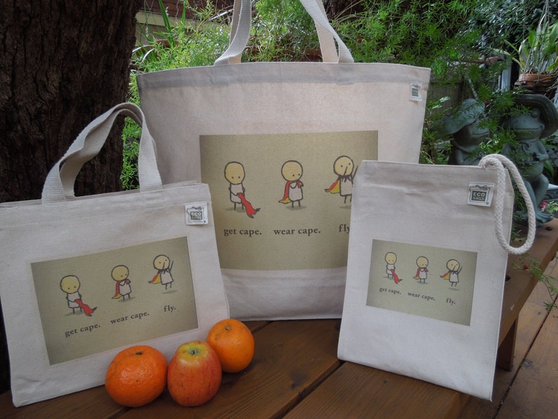 May include: Three white canvas tote bags with colourful illustrations of stick figures wearing capes. The text on the bags reads "get cape. wear cape. fly."