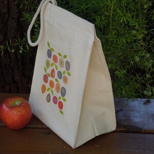 Puede incluir: Una bolsa de almuerzo de lona blanca con un dise&ntilde;o de fruta colorida en la parte delantera. La bolsa tiene un asa de cuerda marr&oacute;n.