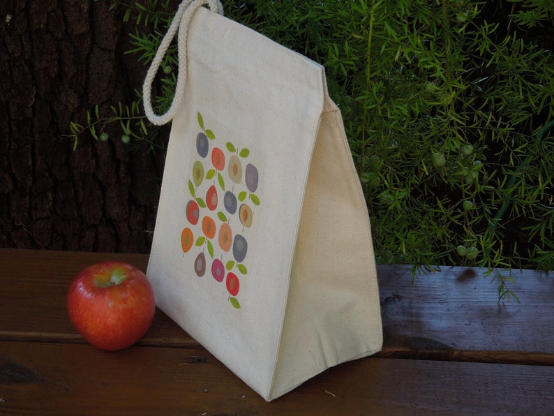 Puede incluir: Una bolsa de almuerzo de lona blanca con un dise&ntilde;o de fruta colorida en la parte delantera. La bolsa tiene un asa de cuerda marr&oacute;n.