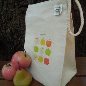 Puede incluir: Una bolsa de lona blanca con asa de cuerda. La bolsa tiene una etiqueta verde que dice "Eco Bags" y un gr&aacute;fico de manzanas en la parte delantera. El texto "Apples" est&aacute; impreso encima del gr&aacute;fico.
