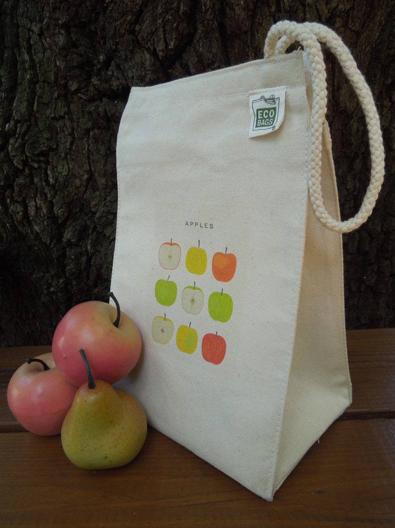 Puede incluir: Una bolsa de lona blanca con asa de cuerda. La bolsa tiene una etiqueta verde que dice "Eco Bags" y un gr&aacute;fico de manzanas en la parte delantera. El texto "Apples" est&aacute; impreso encima del gr&aacute;fico.