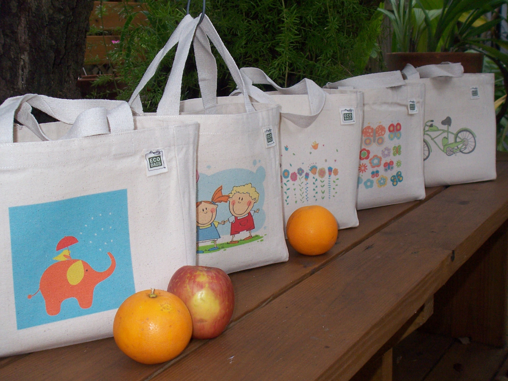 May include: Five white canvas tote bags with different colorful designs. The designs include a floral pattern, a butterfly pattern, a bicycle pattern, a girl and boy pattern, and an elephant pattern. The bags are lined up on a wooden surface.