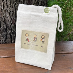May include: A white canvas tote bag with a drawstring closure. The bag has a green logo that says "Est. 1993 ECO BAGS". The front of the bag features a cartoon illustration of three stick figures with the text "get cape. wear cape. fly."