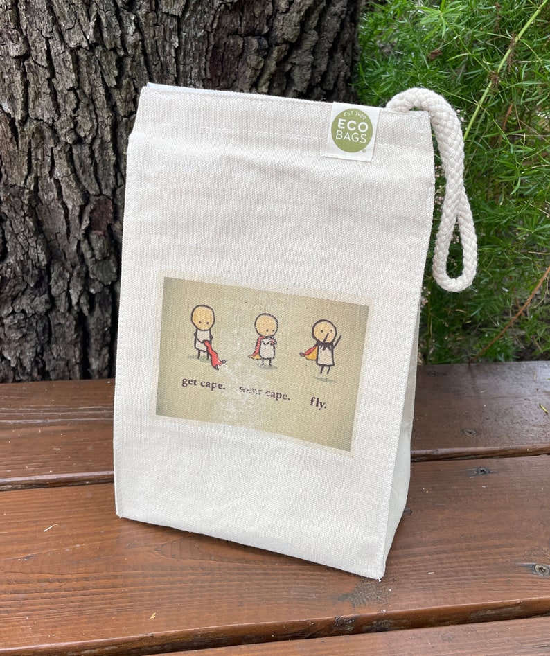 May include: A white canvas tote bag with a drawstring closure. The bag has a green logo that says "Est. 1993 ECO BAGS". The front of the bag features a cartoon illustration of three stick figures with the text "get cape. wear cape. fly."
