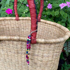 Puede incluir: Una cesta de paja tejida con dos asas resistentes envueltas en cuero rojo con costuras negras. Un cord&oacute;n decorativo de peque&ntilde;as cuentas de madera multicolor en tonos rosa, azul, verde y amarillo cuelga de una de las asas envueltas en cuero.