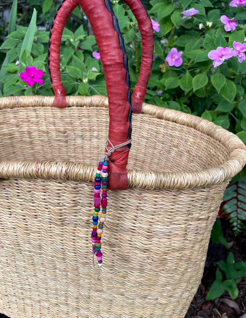 Puede incluir: Una cesta de paja tejida con dos asas resistentes envueltas en cuero rojo con costuras negras. Un cord&oacute;n decorativo de peque&ntilde;as cuentas de madera multicolor en tonos rosa, azul, verde y amarillo cuelga de una de las asas envueltas en cuero.