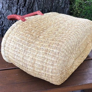 Puede incluir: Una cesta de paja tejida con base rectangular y esquinas redondeadas, que presenta una textura de fibra natural de color tostado claro y un par de asas de cuero marr&oacute;n rojizo cosidas en el borde superior de la cesta.