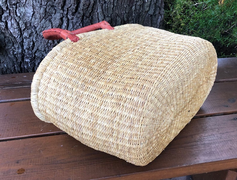 Puede incluir: Una cesta de paja tejida con base rectangular y esquinas redondeadas, que presenta una textura de fibra natural de color tostado claro y un par de asas de cuero marr&oacute;n rojizo cosidas en el borde superior de la cesta.