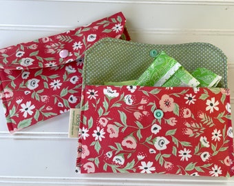Puede incluir: Dos bolsas de tela rojas con estampado floral y detalles verdes. Las bolsas tienen un cierre de solapa con un botón. La bolsa de la derecha está abierta y contiene una compresa higiénica verde. Las bolsas tienen el texto "Bag It" impreso.