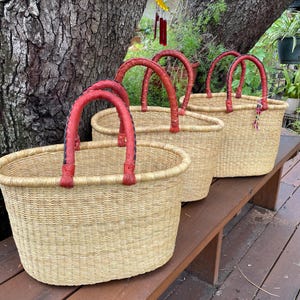Puede incluir: Tres cestas ovaladas tejidas con fibras naturales de color tostado claro. Cada cesta cuenta con dos asas resistentes y redondeadas, envueltas en cuero rojo liso. Las cestas est&aacute;n dispuestas en fila, mostrando sus aberturas y textura.