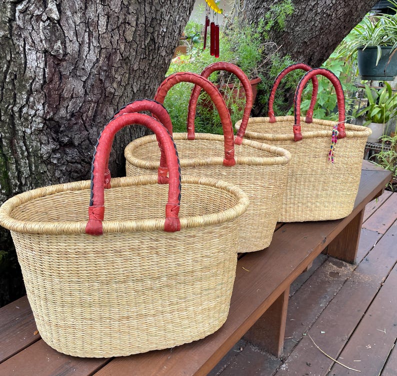 Puede incluir: Tres cestas ovaladas tejidas con fibras naturales de color tostado claro. Cada cesta cuenta con dos asas resistentes y redondeadas, envueltas en cuero rojo liso. Las cestas est&aacute;n dispuestas en fila, mostrando sus aberturas y textura.