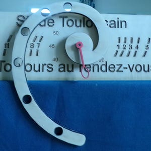 Puede incluir: Un letrero de madera con un diseño en espiral y el texto "de Toulo ain" y "To ours au rendez-vous". El letrero tiene un puntero rojo que se puede mover alrededor de la espiral.