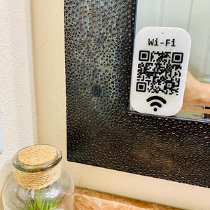 May include: A white Wi-Fi QR code sign with the text "Wi-Fi" and a signal icon. The sign is held in front of a textured black mirror. A glass jar with a cork lid and decorative twine is in the foreground.