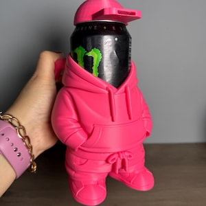 May include: A bright pink 3D-printed beverage can holder shaped like a figure wearing a hoodie, sweatpants, and a baseball cap. The holder contains a black energy drink can with a green logo, and the cap features a small rectangular whistle.