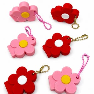 May include: Six flower-shaped keychains in pink and red. Each keychain has a yellow or white center and a small chain. The keychains are made of a soft material and are designed to look like flowers.