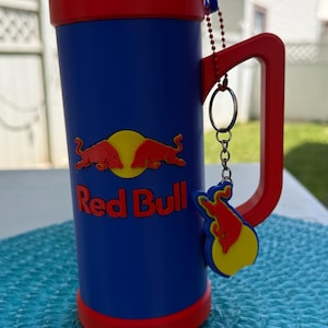 May include: A blue and red plastic can cooler mug featuring the Red Bull logo with two red bulls and a yellow sun. The mug has a red handle with a matching red bull keychain attached by a silver chain. A silver Red Bull energy drink can is inside.