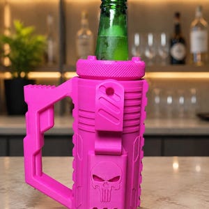 May include: A bright pink, cylindrical bottle holder with a textured handle and a skull emblem embossed on the front. The holder features a screw-top rim and a vertical clip, currently containing a green glass bottle with a narrow neck.