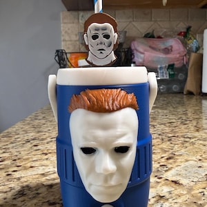 May include: A blue beverage cooler featuring a 3D-printed white mask with brown hair on the front. The cooler has a white lid, a white spigot at the base, and a blue and white striped straw topped with a small matching mask decoration.