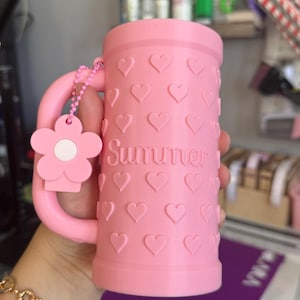May include: A cylindrical pink mug featuring a repeating embossed heart pattern across the exterior surface. The word Summer is embossed in the center. A pink flower-shaped charm hangs from the handle by a small pink beaded chain.