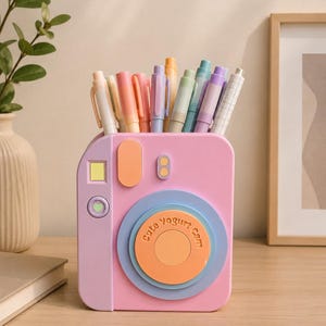 Puede incluir: Un organizador de escritorio rosa con forma de cámara lleno de bolígrafos de colores pastel. El frente presenta un diseño de lente circular con el texto "Cute Yogurt Cam" impreso en naranja, junto a un cuadrado amarillo, un óvalo naranja y un pequeño corazón.