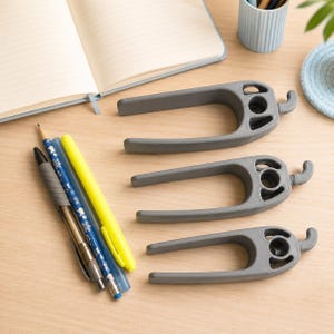 May include: Three gray, U-shaped plastic desk organizers with a small hook on one end and a circular opening on the other. These items are positioned next to a collection of writing instruments, including a yellow highlighter and two blue pens.