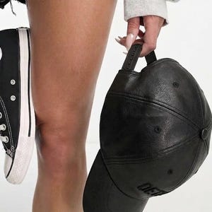 K&ouml;nnte beinhalten: Schwarze Baseballkappe mit gebogenem Schirm und verstellbarem Riemen. Die Kappe hat eine strukturierte Optik und das Wort "DE" ist auf der Vorderseite zu sehen. Ein schwarzer und wei&szlig;er Canvas-Sneaker ist ebenfalls im Bild.