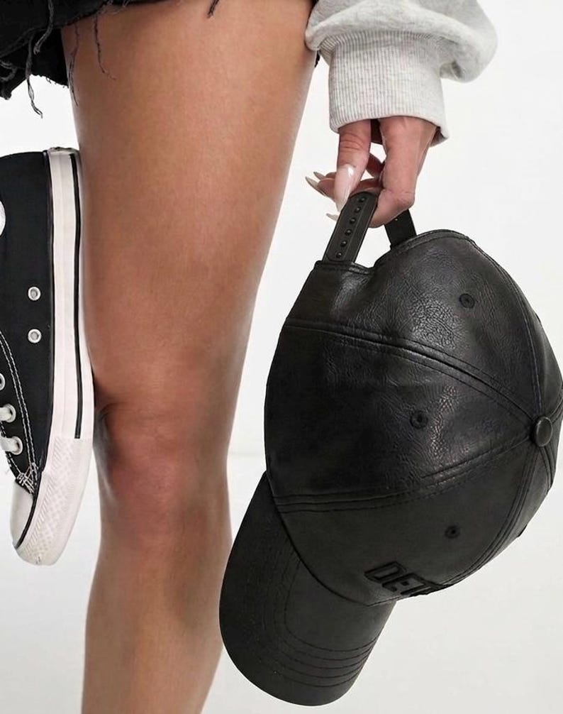 K&ouml;nnte beinhalten: Schwarze Baseballkappe mit gebogenem Schirm und verstellbarem Riemen. Die Kappe hat eine strukturierte Optik und das Wort "DE" ist auf der Vorderseite zu sehen. Ein schwarzer und wei&szlig;er Canvas-Sneaker ist ebenfalls im Bild.
