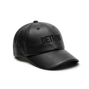 K&ouml;nnte beinhalten: Schwarze Baseballkappe mit dem Wort "DETROIT" auf der Vorderseite. Die Kappe hat einen gebogenen Schirm und einen Knopf oben. Der Hut ist aus einem glatten, lederartigen Material gefertigt.