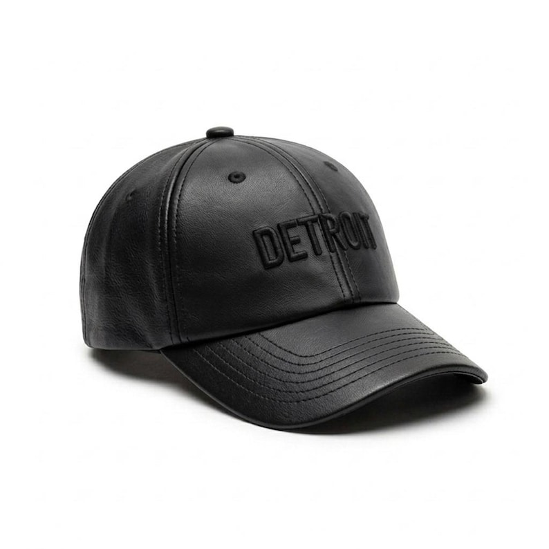 K&ouml;nnte beinhalten: Schwarze Baseballkappe mit dem Wort "DETROIT" auf der Vorderseite. Die Kappe hat einen gebogenen Schirm und einen Knopf oben. Der Hut ist aus einem glatten, lederartigen Material gefertigt.