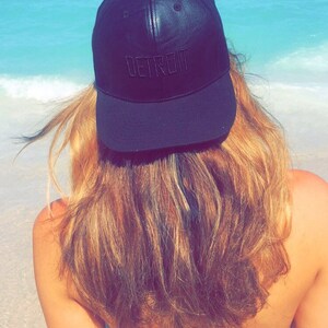 K&ouml;nnte beinhalten: Eine schwarze Baseballkappe mit dem Wort "DETROIT" auf der Vorderseite gestickt. Die Kappe wird von einer Person mit langen, hellbraunen Haaren getragen, mit einem Strand und Meer im Hintergrund.