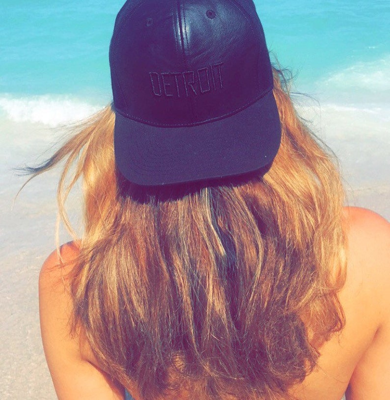 K&ouml;nnte beinhalten: Eine schwarze Baseballkappe mit dem Wort "DETROIT" auf der Vorderseite gestickt. Die Kappe wird von einer Person mit langen, hellbraunen Haaren getragen, mit einem Strand und Meer im Hintergrund.