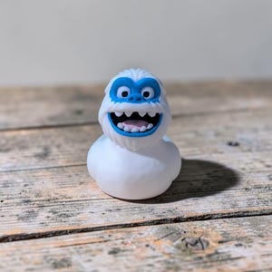 May include: A white rubber duck shaped like a yeti with a blue face mask, large round eyes, and a wide open mouth showing white teeth and a pink tongue. The figure has a smooth, rounded body and a textured head resembling thick fur or hair.