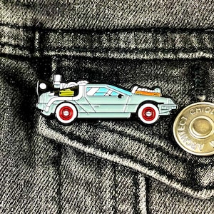 May include: A silver and blue enamel pin of the DeLorean time machine from the Back to the Future movie. The pin is on a denim jacket.