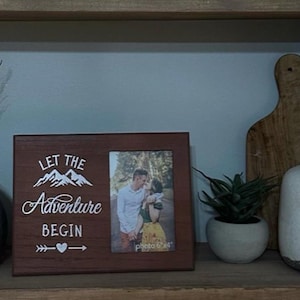 May include: A brown wooden picture frame with a white inscription that reads "Let the Adventure Begin" and a heart with an arrow. The frame contains a photo of a couple in a forest setting.
