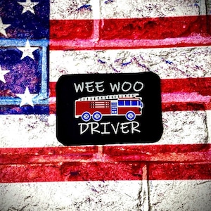 May include: A black patch with the words "WEE WOO DRIVER" and a red and blue fire truck graphic. The patch is set against a backdrop of the American flag, painted on a brick wall.