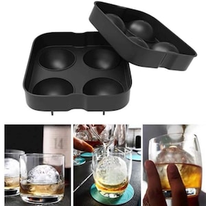May include: Black silicone ice mold with four round cavities for making large ice spheres. The mold is open and shows the ice spheres in a glass of whiskey.
