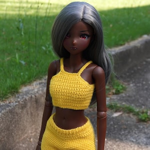 May include: A doll with dark skin and gray hair wearing a yellow crochet crop top and matching shorts. The doll is standing on a concrete surface with green grass in the background. The doll has red eyes.