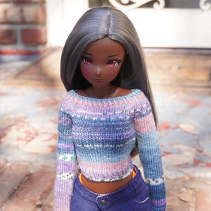 Könnte beinhalten: Eine Puppe mit dunkler Haut und grauem Haar trägt einen gecroppten, gestreiften Pullover in Blau-, Lila- und Rosatönen. Die Puppe trägt auch blaue Jeansshorts. Die Puppe hat rote Augen und steht draußen.