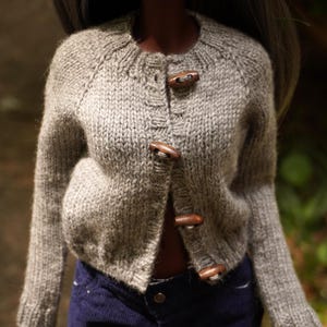 May include: A gray knitted cardigan with wooden toggle closures. The sweater has a round neckline and long sleeves. The cardigan is worn over a dark blue garment.