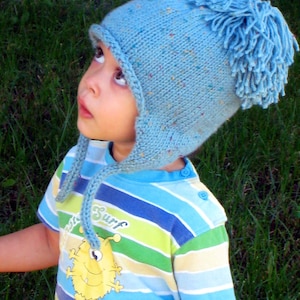 May include: A child wearing a blue knitted hat with a large blue pom-pom on top. The hat has ear flaps and a long tie that hangs down the front of the child's shirt. The child is wearing a blue, green, and yellow striped shirt with a cartoon monster graphic that says "Surf".
