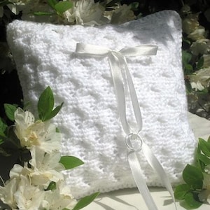 Puede incluir: Almohada de crochet blanco con una cinta de satén y dos anillos plateados. La almohada está decorada con un delicado patrón de encaje.