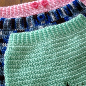 May include: Three crocheted shorts in pink, blue and green with white and brown accents. The shorts have buttons on the waistband.