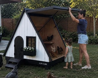 Könnte beinhalten: A-förmiger Hühnerstall mit weißem Äußeren und schwarzen Zierleisten. Der Stall hat eine kleine Tür, einen Blumenkasten und eine Rampe. Hühner sind im und außerhalb des Stalls. Eine Frau und ein Kind sind in der Nähe des Stalls.