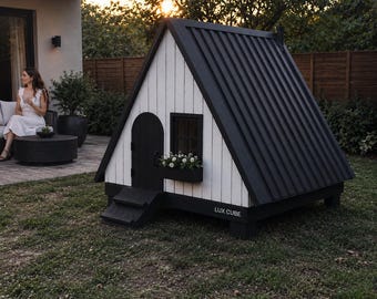 Könnte beinhalten: Ein A-förmiges Hundehäuschen mit schwarzem Metalldach und schwarzem Zierrat. Das Haus hat eine weiße Außenseite mit einer kleinen Tür und einem Fenster. Unter dem Fenster befindet sich ein Blumenkasten mit weißen Blumen. Auf der Basis steht "LUX CUBE".
