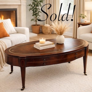 May include: An oval wooden coffee table with a dark brown finish, featuring a gold-toned decorative border along the edge, two small drawers with dark knobs, and four tapered legs with metal casters. The word Sold! is written in black script.