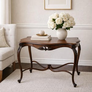 May include: A dark brown, antique-style side table with ornate carved details and curved legs. A white vase filled with white roses sits on the table, along with books and a decorative sphere. The table is placed on a light-colored rug.