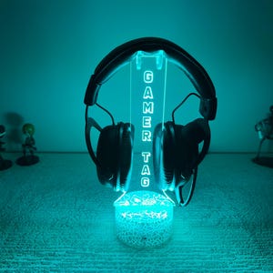 May include: A black gaming headset rests on a teal-colored illuminated stand. The stand displays the text "GAMER TAG" in a vertical font. The base of the stand has a decorative, openwork design. The background is a solid teal color.